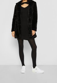 Manteau en fausse fourrure noire, robe noire à côtes avec un design découpé, collants noirs et baskets blanches complètent la tenue.