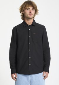 Chemise noire à boutons avec une seule poche poitrine, boutons blancs et un petit logo orange sur la gauche. Fabriquée en tissu lisse et léger.