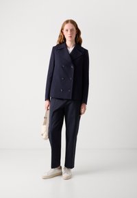 Veste bleu marine croisée avec de grands boutons noirs, associée à un pantalon sur mesure assorti et des baskets blanches. Le modèle tient un sac à main beige.