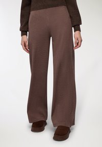 Brązowe spodnie typu wide-leg z teksturowanym wzorem w kratkę, wykonane z miękkiego materiału. Połączone z ciemnobrązowymi butami na platformie.