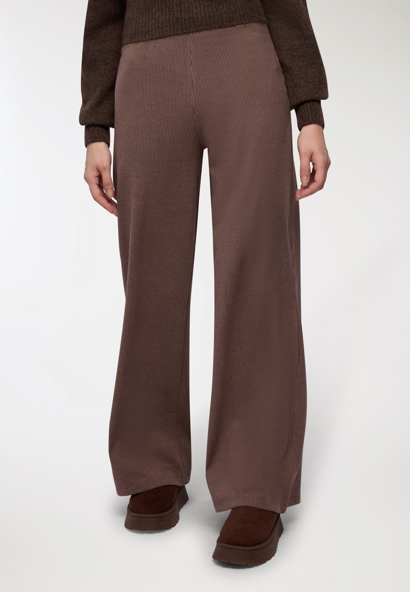 Brązowe spodnie typu wide-leg z teksturowanym wzorem w kratkę, wykonane z miękkiego materiału. Połączone z ciemnobrązowymi butami na platformie.