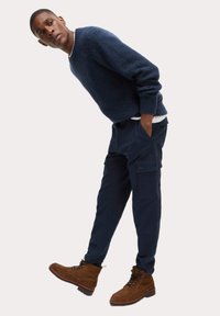 Marine gebreide trui, marine cargobroek met zijzakken, en bruine veterschoenen. De outfit is casual en functioneel met een relaxte pasvorm.