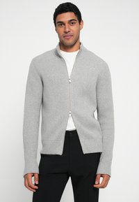 Cardigã cinza canelado com fecho de correr, gola alta, mangas compridas e bainha reta. Apresenta bolsos laterais e fecho de correr liso.