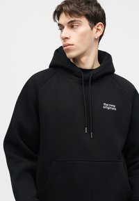 Mladi moški v črni kapucarji z napisom "the new originals", gleda levo, na svetli enobarvni podlagi.