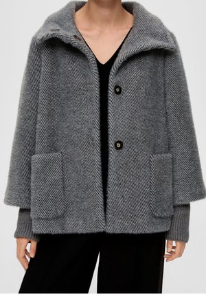 Femme portant un manteau en laine texturée gris avec de grandes poches, un large col et des boutons noirs sur un haut et un pantalon noirs.