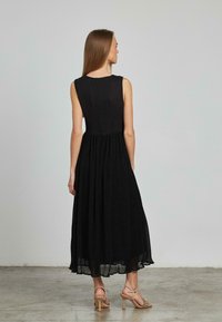 Schwarzes ärmelloses Kleid mit einer figurbetonten Oberseite und einem fließenden, plissierten Rock. Reißverschlussdetail hinten; getragen mit beigen, hohen Sandalen mit Riemchen.