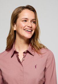 Camicia button-up rosa con colletto classico, due bottoni al collo e un piccolo logo ricamato sul petto sinistro. Texture liscia.
