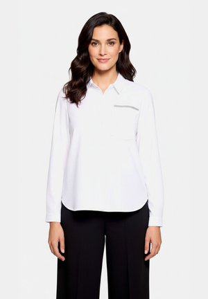 Femme aux cheveux foncés portant une chemise blanche à manches longues avec une poche poitrine et un pantalon noir, debout devant un fond clair uni.