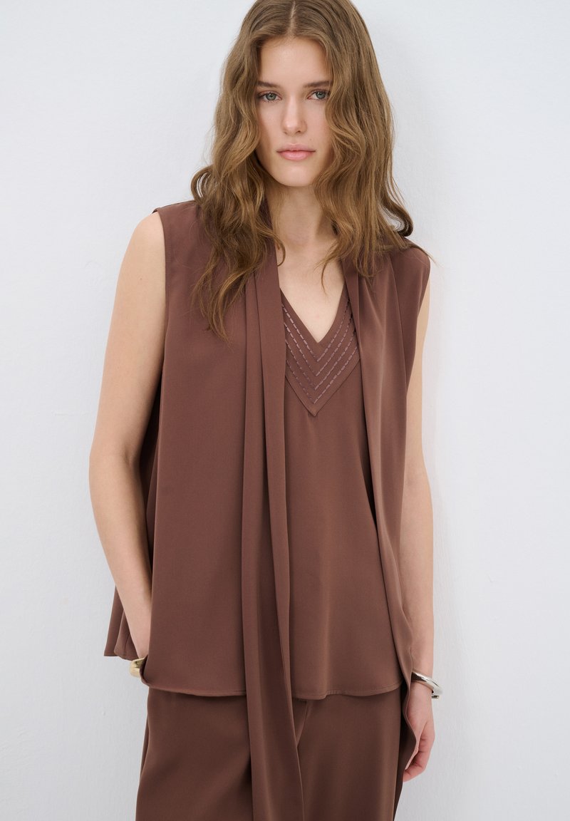 Bruine mouwloze blouse met een V-hals, voorzien van een gedrapeerd ontwerp en gestikte details bij de halslijn. Zachte, vloeiende textuur van de stof.