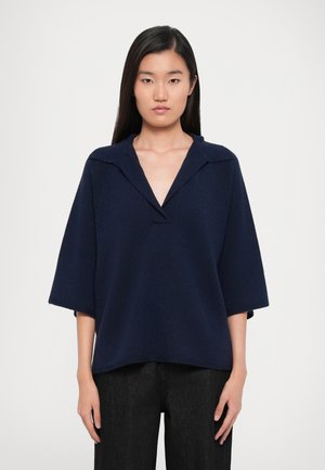 Maglione over-size blu navy con scollo a V, maniche corte e ampie, e un motivo a maglia testurizzato, abbinato a pantaloni in denim scuri.