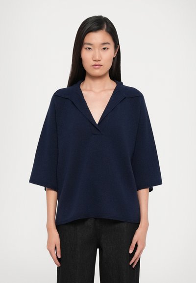 Suéter azul marino de gran tamaño con escote en V, mangas cortas anchas y un patrón de punto texturizado, combinado con pantalones de mezclilla oscuros.