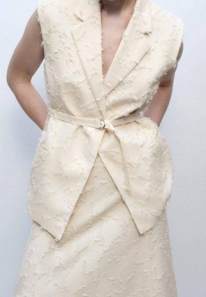 Ærmeløs beige tekstureret blazer med matchende nederdel, båret med bælte i taljen og hænderne i lommerne.