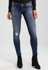 Jeans ajustados de mezclilla azul oscuro con detalles desgastados, que cuentan con una cintura de altura media y una silueta ajustada. Combinados con botas negras hasta el tobillo.