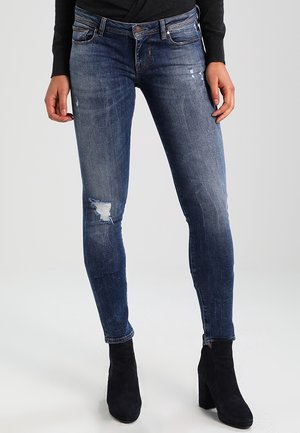 Jeans ajustados de mezclilla azul oscuro con detalles desgastados, que cuentan con una cintura de altura media y una silueta ajustada. Combinados con botas negras hasta el tobillo.