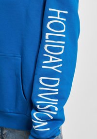 Sudadera azul con un bolsillo delantero y texto blanco que dice "HOLIDAY DIVISION" en vertical a lo largo de la manga izquierda. Tejido suave y texturizado.