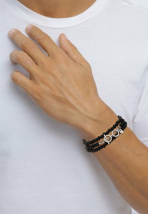 Hand mit gebräunter Haut ruht auf der Brust, trägt ein schwarzes Perlenarmband mit silbernen Verschlüssen über einem schlichten weißen Hemd.