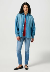 Camisa de mezclilla en azul claro con botones en la parte delantera, mangas largas y corte relajado, sobre una camiseta roja. Combinada con jeans azul claro y zapatos planos negros.