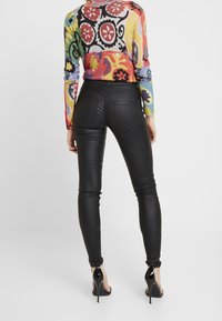 Vrouw in zwarte leren broek, een kleurrijke bloemenpatroon langemouwtop en zwarte hoge hakken, staand met de rug naar ons gekeerd.