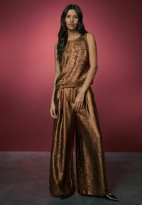 Bronze metallic sleeveless top with matching wide-leg pants, featuring a textured finish and relaxed fit, set against a pink background.