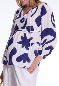 Witte blouse met marineblauwe abstracte patronen, lichtgewicht gekreukt materiaal, trekkoord-halslijn en lange pofmouwen, gecombineerd met een witte brede broek.