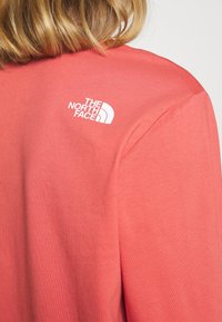 Korallinvärinen t-paita, joka on valmistettu sileästä ja kevyestä kankaasta. Vasemmassa olkapäässä sijaitsee valkoinen logo. Lyhyet hihnat ja rento malli.