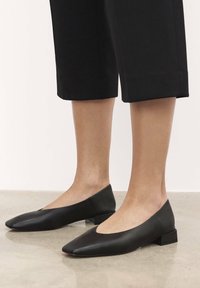 Balerini negri din piele cu vârf ascuțit, toc mic pătrat și design cu decupaj pe laterale. Textură netedă cu o estetică minimalistă.