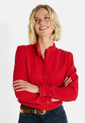 Blouse rouge à côtes avec une texture douce, col volanté et fermeture à boutons. Manches longues avec boutons aux poignets. Portée avec une ceinture marron.