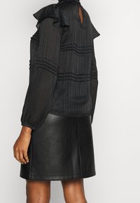Blouse noire plissée avec des épaules volantées et un dos ajouré ; associée à une jupe en cuir noire, mettant en valeur une texture lisse et un design élégant.