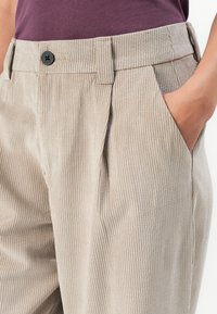 Beige gerippte Cordhosen mit einem schwarzen Knopf und Gürtelschlaufen, getragen mit einem in die Hose gesteckten dunkelvioletten Oberteil, Hand in rechter Tasche.