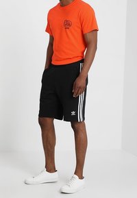 T-shirt en coton orange avec impression graphique, associé à un short noir avec des rayures blanches sur les côtés. Complété par des baskets blanches.