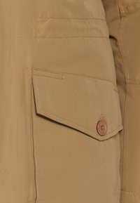 Lichtgewicht stof in tan met een gestructureerde afwerking, voorzien van een klepzak met een ronde bruine knoopdetails. Eenvoudig, utilitair ontwerp.