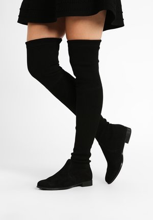 Benen met zwarte over-de-knie platte laarzen gecombineerd met een zwarte rok, staand met één been gekruist voor het andere.