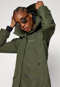 Grüne wasserdichte Jacke mit hohem Kragen, Frontreißverschlüssen und verstellbarem Kapuzen. Mit Logo auf der Brust und texturiertem Stoff.