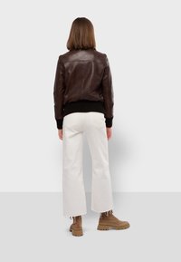 Braune Lederbomberjacke mit gerippten schwarzen Bündchen und Saum. Kombiniert mit weißen weit geschnittenen Jeans und braunen klobigen Stiefeln. Ansicht von hinten.