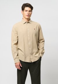 Cămașă bej cu mânecă lungă, cu închidere prin buton, guler ascuțit și un mic accent brodat. Purtați cu pantaloni verzi închis; material texturat.