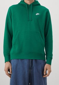 Vihreä puuvillainen huppari, jossa on kengurutasku, kiristettävä huppu ja valkoinen Nike-logo rinnassa. Varustettu resorikämmensuuilla ja helmalla.