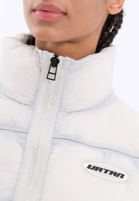 Ljusgrå pufferjacka med hög krage, försedd med en framdragkedja, svart detaljer och en logotyp-patch med svart text på bröstet.
