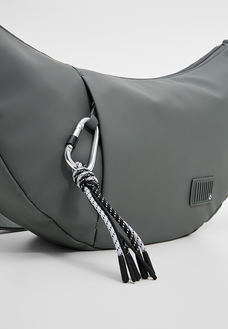 Bolsa de nailon gris con forma curvada, que presenta un mosquetón plateado, acentos de cuerda en blanco y negro, y un parche de logo texturizado.