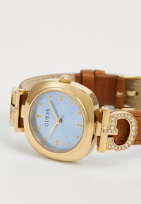 Goldfarbene Guess-Armbanduhr mit hellblauem Ziffernblatt, Kristall-Stundenmarkierungen und braunem Lederarmband mit mit Kristallen besetzter Schnalle.