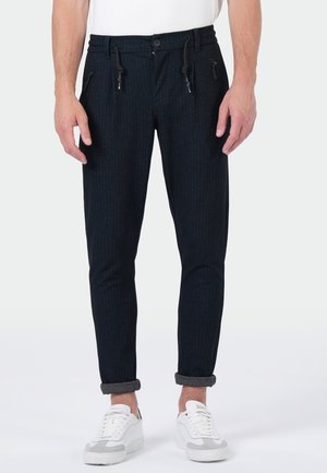 Man draagt opgerolde zwarte broek met fijne strepen, trekkoord en ritszakken, gecombineerd met witte sneakers op een witte achtergrond.