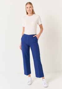 Weißes gestricktes Oberteil mit kurzen, rüschengesäumten Ärmeln, kombiniert mit blauen weit geschnittenen Hosen mit vorderen Taschen und Knopfdetails, getragen mit weißen Sneakers.