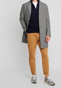 Houndstooth-mönstrad kappa i grått och svart, mörk marinblå tröja, beige chinos och grå sneakers med vita snören. Avslappnad passform och lagrad stil.