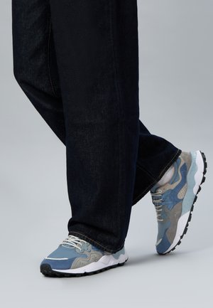 Persona che indossa scarpe da ginnastica blu e grigie con suole bianche e jeans scuri su uno sfondo grigio uniforme.