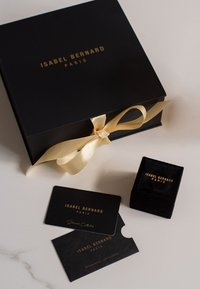 Boîte-cadeau noire avec du texte doré et un ruban crème. Comprend une boîte à anneau en velours noir et deux cartes noires avec une inscription dorée.