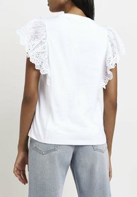 Vit bomull t-shirt med ögla spets volangärmar. Bakgrunden visar en enkel design med rak fåll och mjuk textur. Bärs med ljusblå jeans.