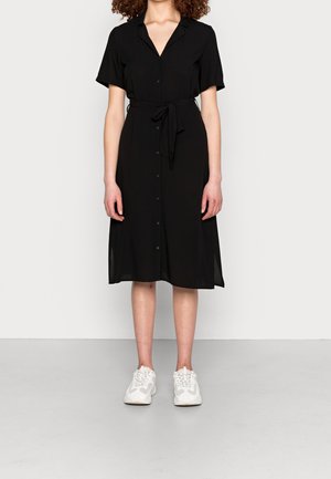 Kobieta ubrana w czarną sukienkę midi na guziki z krótkimi rękawami i związaną talią, w połączeniu z białymi sportowymi sneakersami, stojąca na szarej podłodze.