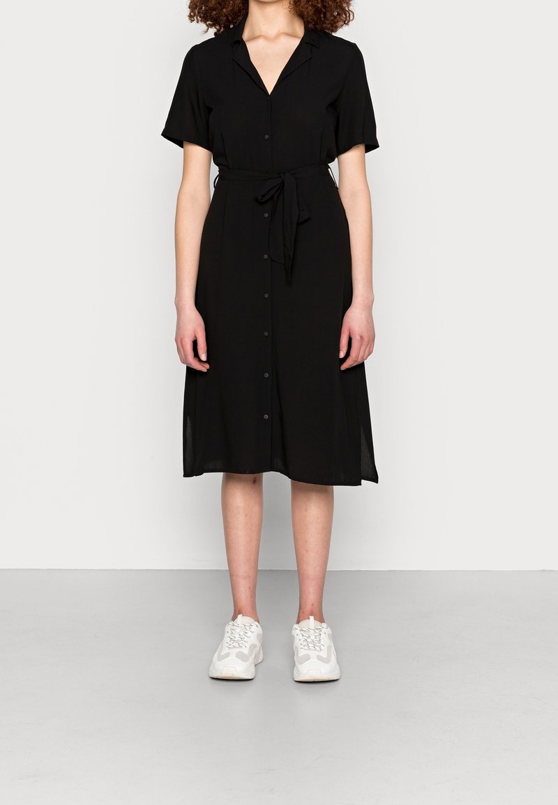 Femme portant une robe midi noire boutonnée, à manches courtes et taille nouée, associée à des baskets blanches de sport, debout sur un sol gris.