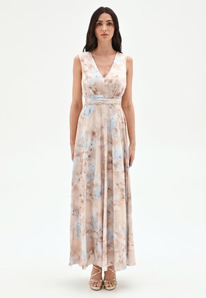 Donna con capelli lunghi e scuri che indossa un vestito beige senza maniche, con scollo a V, lungo fino al pavimento, con motivi floreali blu e marroni e scarpe dorate con tacchi alti.