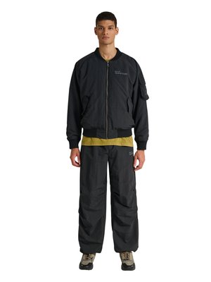 Jonge man staand, draagt een zwarte bomberjack met rits, losse zwarte broek, gele shirt eronder en beige wandelschoenen op een witte achtergrond.