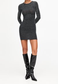 Robe mini noire à manches longues avec une silhouette ajustée, fabriquée à partir d'un tissu texturé présentant un subtil scintillement. Des bottes noires montantes complètent le look.